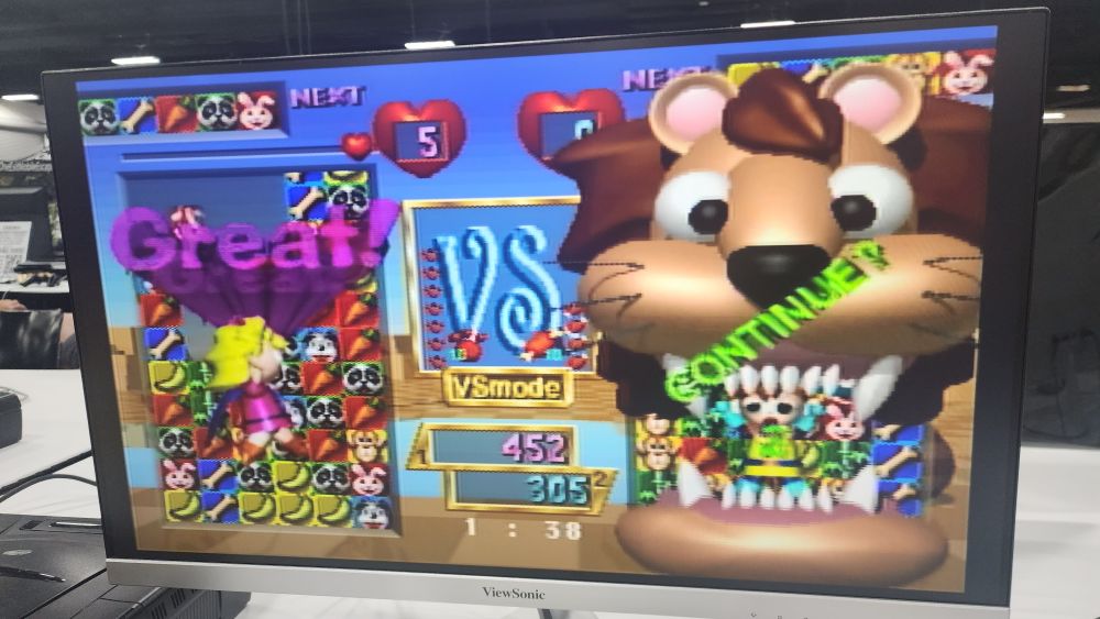 Photo of an LCD TV playing Baku Baku Animal. The game is on a victory screen from a VS battle. The graphics are surreal 3D models with harsh lighting. A giant lion head engulfs the right playfield and a dancing girl is on the left playfield.