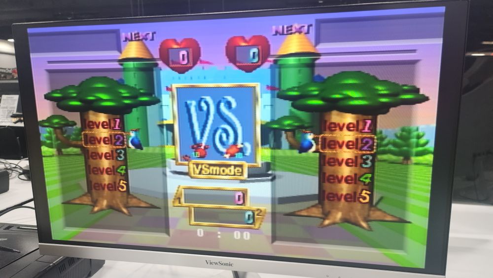 Photo of an LCD TV playing Baku Baku Animal. The game is on a level select. The graphics are surreal 3D models with harsh lighting