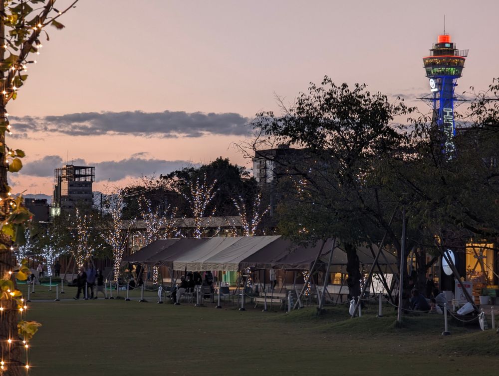 天王寺公園 の写真
夕暮れの空の下、公園または広場で開かれているイベントの様子。右奥には時計と通天閣が見える。手前の芝生の上には複数のテントが並び、周囲の木々はイルミネーションで飾られている。左端の木にもイルミネーションが施されている。