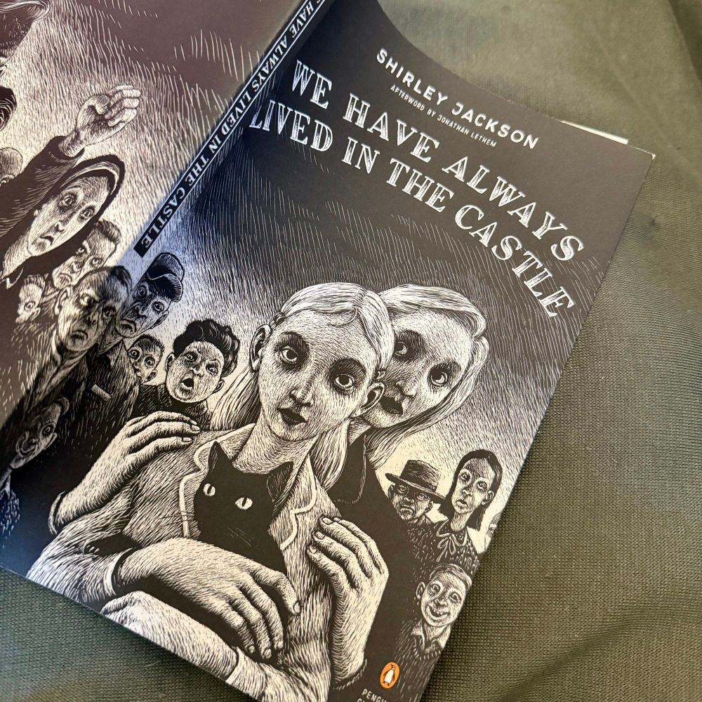 Paperback edition of We Have Always Lived in the Castle by Shirley Jackson. The book is open and face down on a green blanket. The cover is illustrated in black and white and shows a teenage (?) girl holding a black cat with a woman standing behind her. The woman hands are on the teenagers shoulders and behind them is an illustrated crowd of people with a variety of facial expressions. 
