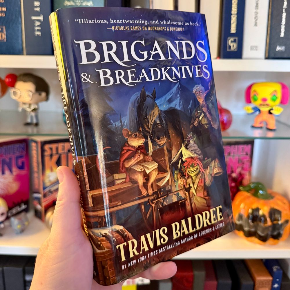Brigands & Breadknives by Travis Baldree. The cover shows a traveling trio camped out around a fire and a cookpot. There's a mouse with a red cape sitting on the back of a wagon, writing, and a draft horse is standing next to the mouse’s shoulder. Next to the cookpot appears to be a little green goblin with an oversized patchwork jacket and a pointed hat covering bright red hair, red eyes, and pointed teeth. Standing behind the goblin, slightly in shadow, and next to the horse's hindquarters is an annoyed-looking elf with its sword partially drawn from the sheath. 