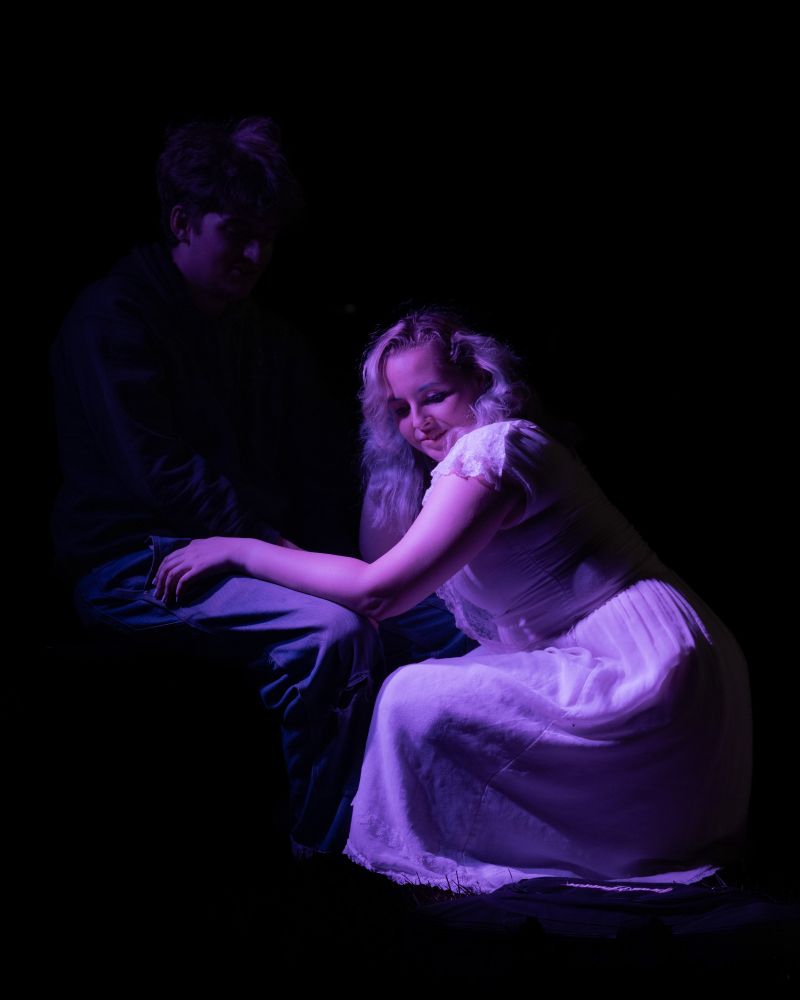 A woman in a white dress, face turned towards the camera, kneels in from on a man largely hidden from view.
