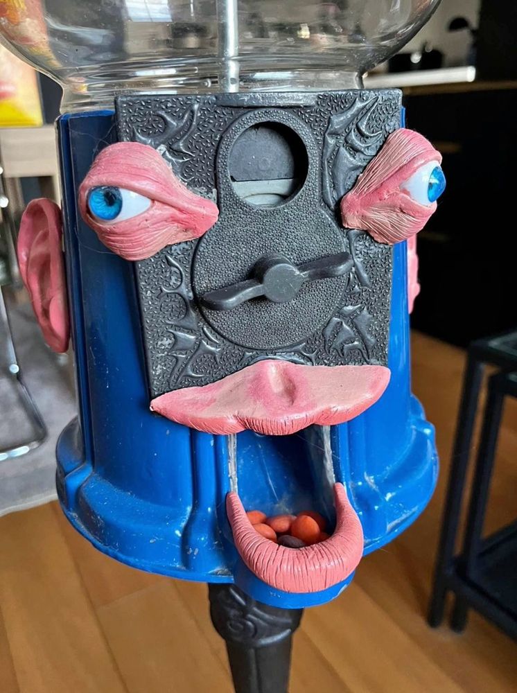 An old fashioned gumball machine with the body of the machine decorated to have very lifelike sculpted ears and blue bulging wall-eyes, with the opening where gumballs come out turned into a mouth with realistic upper and lower lips.