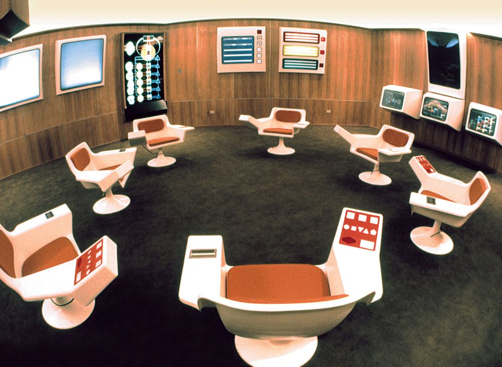 a ring of seven cybernetics chairs in a room with an early 70's sci-fi corporate decor 