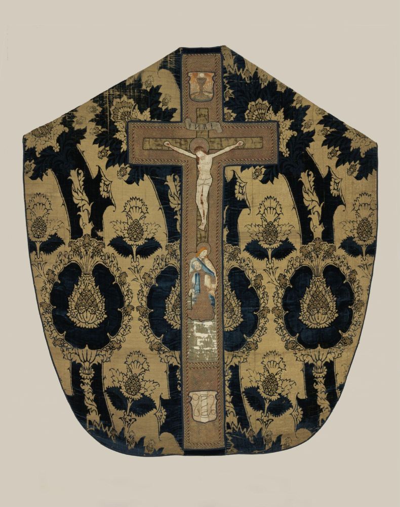 A chasubule, which a priest would wear, of cloth of gold, featuring a crucifix and Mary below it. The velvet behind it is deep indigo in color.