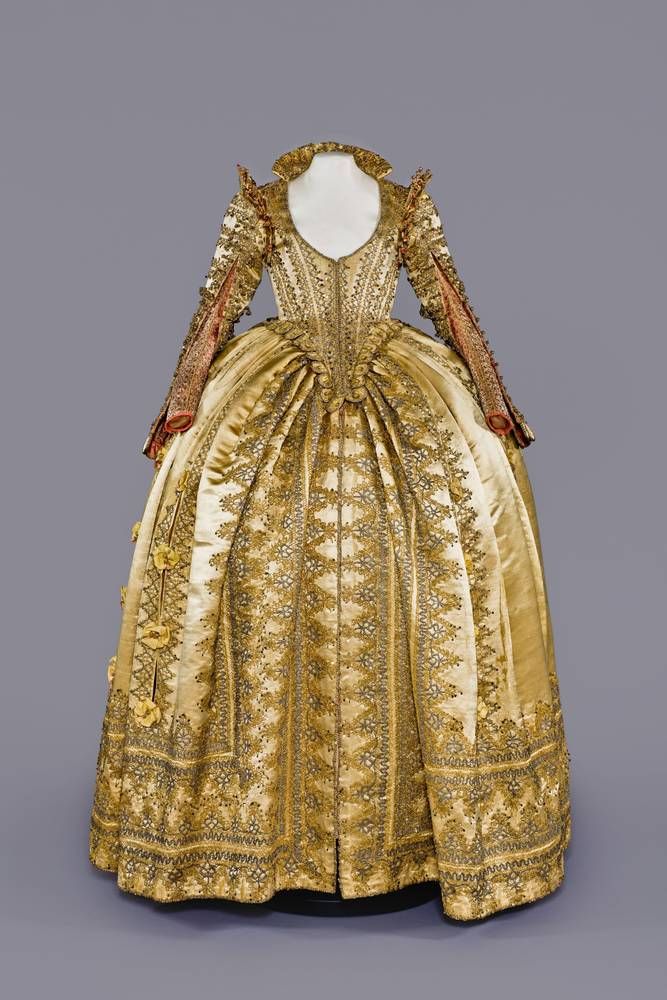 From State Art Collection - Dresden - The golden yellow and salmon-red ceremonial dress with gold and silver lace follows the shape, nuanced colors and sophisticated decorative details of Italian women's fashion around 1590-1620.
The tight-fitting, slightly protruding, pointed bodice is trimmed in dense rows with narrow gold and silver lace. It is closed at the front with concealed hooks and eyes. The skirt is buttoned to the bodice using numerous gold and silver trimming buttons. The slightly stiffened bodice embodies an early form of the so-called snail waist, which was to be a defining feature of women's fashion in the 17th century.
The skirt - an overskirt - is open at the front and is effectively trimmed in several circumferential panels with wide and medium-wide gold and silver lace, into which drop-shaped silver sequins are worked. A second, representative skirt was effectively visible through the front slit and the long skirt slits, which were held together with ribbon roses (not known).
The graceful star-shaped shoulder decoration is borrowed from the bridal and formal wear of noble Venetian women. The jagged tips flash with bright red silk pompoms. The tight-fitting salmon-red sleeves are fastened at the wrist with buttons. They are completely embroidered with scales and stars. The golden yellow wing sleeves above are decorated with lace and many small trimmings made of gold and silver thread.
The accessories of the dress can be imagined as a high, fine white lace collar, lace cuffs of the same kind, and rich gold and pearl jewelry.
The queens and princesses of the English and Danish royal families, as well as Maria de' Medici, Duchess of Florence and Queen of France, were particularly pioneers of the fashion represented by the dress shown. No other original examples have survived in this completeness and condition.