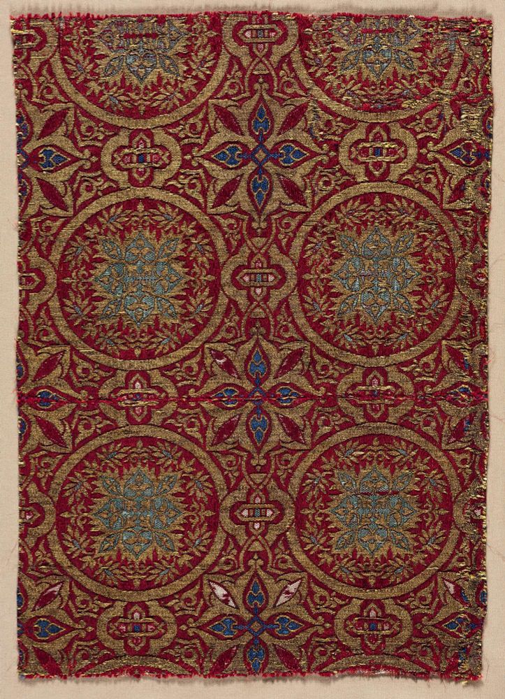 An ornate piece of cloth with roundels in blue and gold against a deep red background. There are also flower-like and cross-like motifs in the background. 