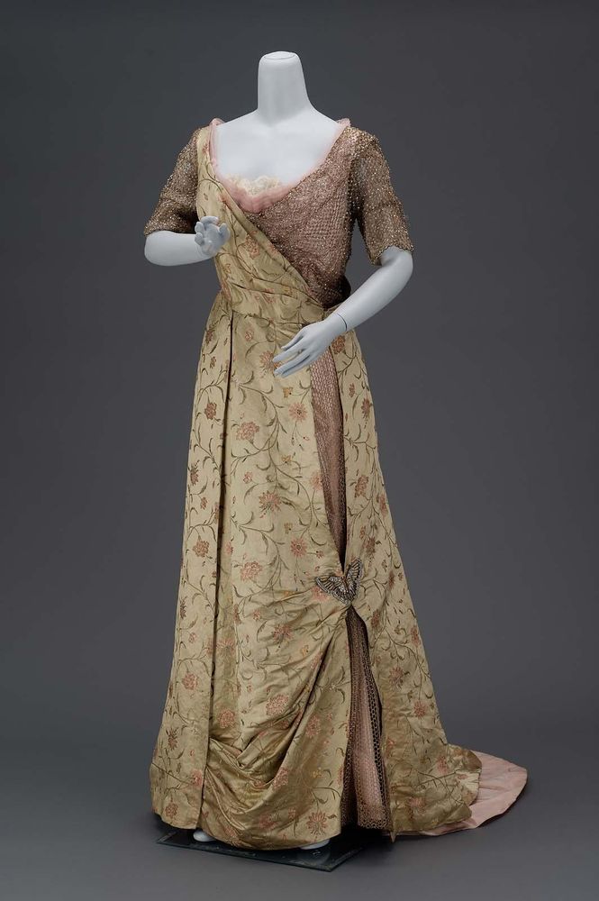 An evening dress of green satin brocaded with pink, green, yellow, and blue silks, and gold metal thread in all-over design of serpentine stems bearing a variety of carnation blossoms; low cut neck with surplice line both front and back of bodice, hooked down back of bodice, short straight sleeves, skirt without much fullness, longer in back forming train,open down entire length of left side to show gold fish net over pink taffeta, this opening caught at center by butterfly of rhinestones, left side of bodice and both sleeves made of gold fish net with rhinestones at intersections of netting.