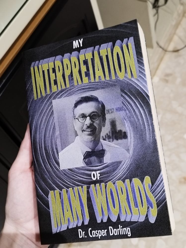 Fotografía de un libro falso llamado "My Interpretation of Many Worlds" escrito por el personaje Casper Darling de los videojuegos Control y Alan Wake 2. Se ve una imagen de Darling en el centro de la portada, rodeada por una espiral gris y el título del libro en letras tridimensionales amarillas.