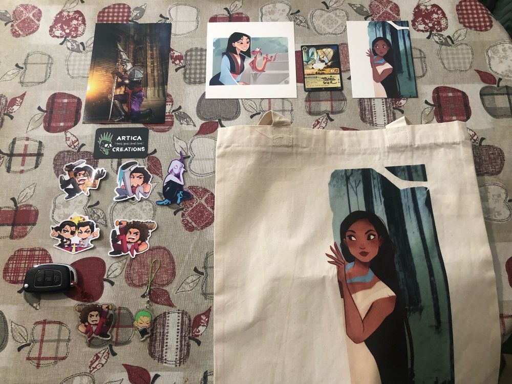 Foto de una mesa con varios objetos de artesanía comprados en FicZone Granada. Se ven varias láminas de arte de Pocahontas y Mulán, pegatinas de la saga Yakuza, llaveros, una tote bag con un diseño de Pocahontas. 