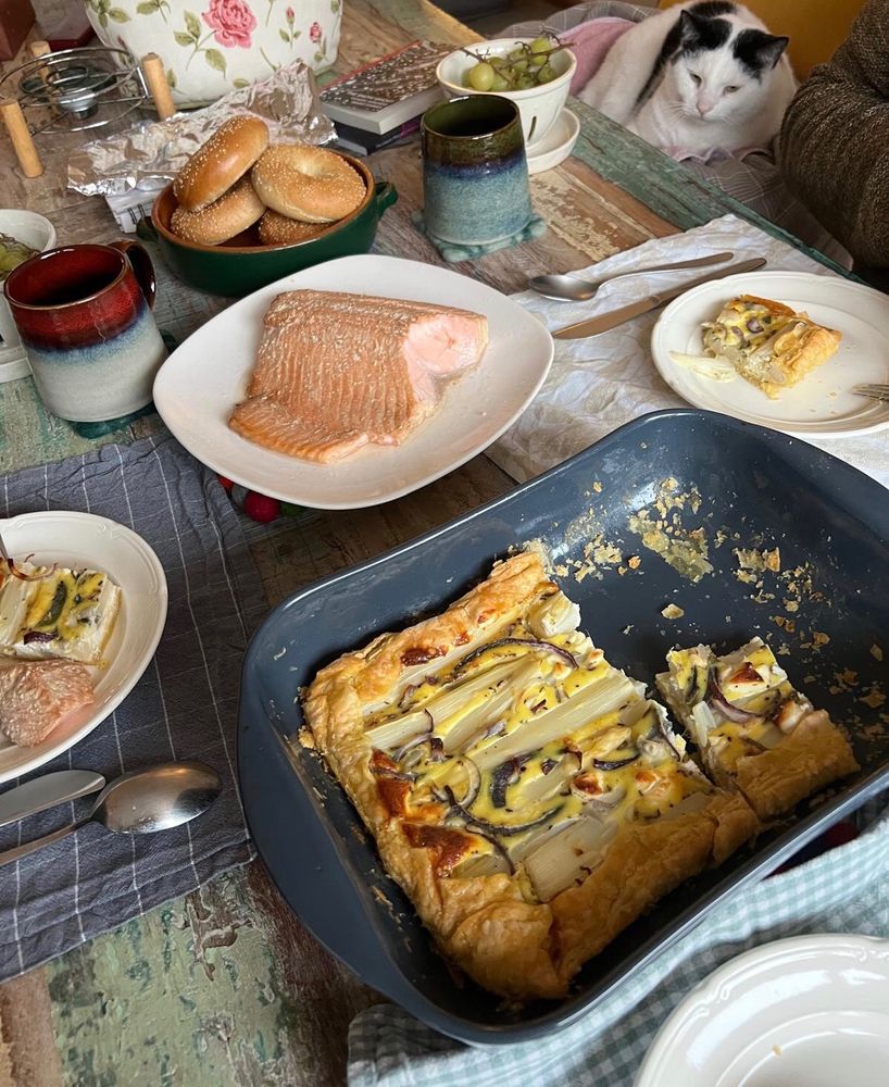 Photo of a brunch table with asparagus quiche, a piece of home-smoked salmon, and bagels. A white cat in the corner of the picture, eyeing the spread from his chair. 