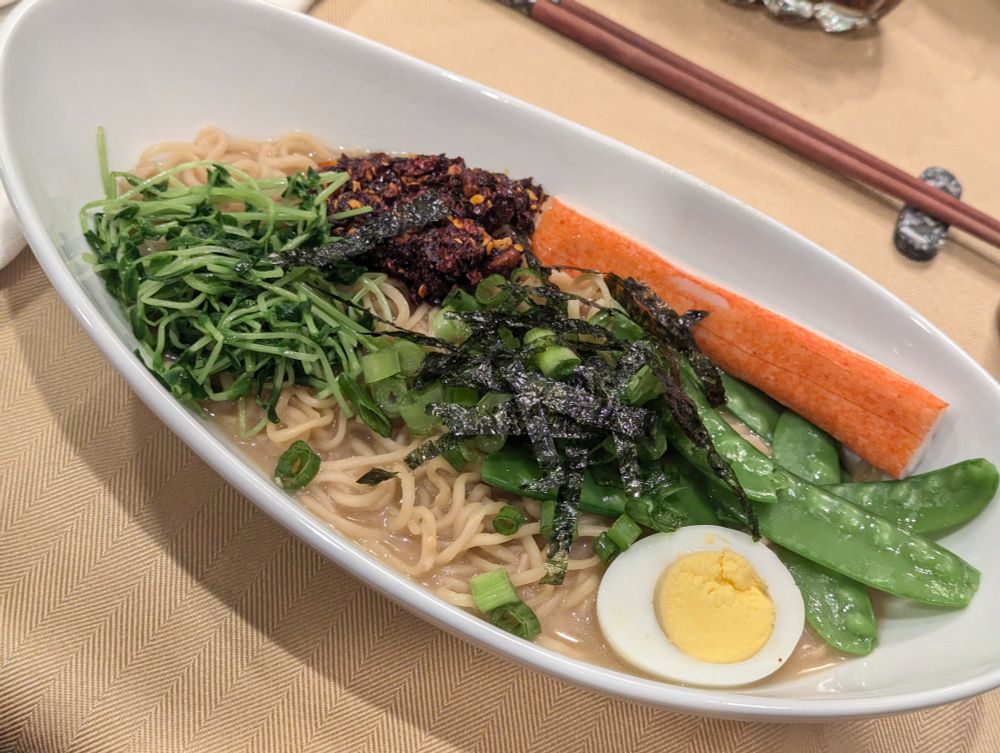 An oblong bowl of ramen noodles and broth with several toppings on top of the noodles.  The toppings are grouped by type. From the leftmost, rotating clockwise, they are pea shoots, chili crisp, crab stick, snow peas, half a boiled egg, and sliced green onions in the center with thin strips of nori placed on top.

The tablecloth is plain, and there are chopsticks on a rest in the background.