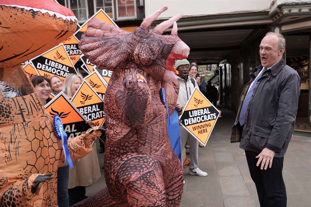 Sir Ed Davey standomg laughing at someone dressed up as a dinosaur, on the campaign trail with the Lib Dems. Despite describing the Tories as dinosaurs, Davey was happy enough to serve under them as Parliamentary Under-Secretary of State for Employment Relations, Consumer and Postal Affairs from 2010 to 2012, and in David Cameron's Cabinet as Secretary of State for Energy and Climate Change from 2012 to 2015.