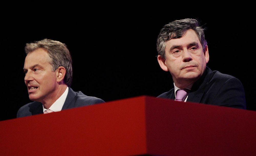 Tony Blair & Gordon Brown looking quite tense at a table at Labour Party Conference. Blair is pulling an awkward smile, while Brown is looking away with a miffed face.