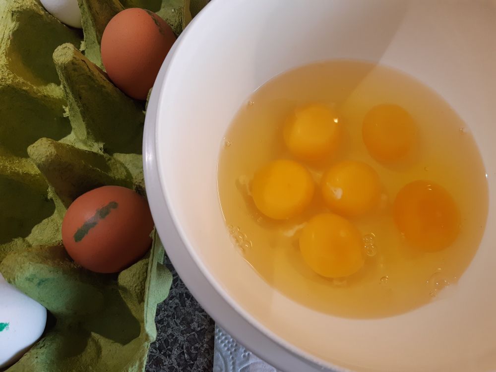 Eine Schüssel mit sechs aufgeschlagen Eiern. Daneben ein grüner Eierkarton mit vier weiteren Eiern.