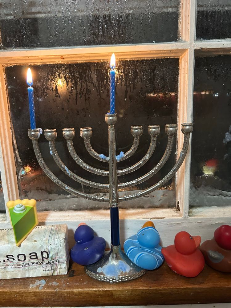A menorah with two lit candles. It’s sitting on a windowsill with some rubber ducks. 