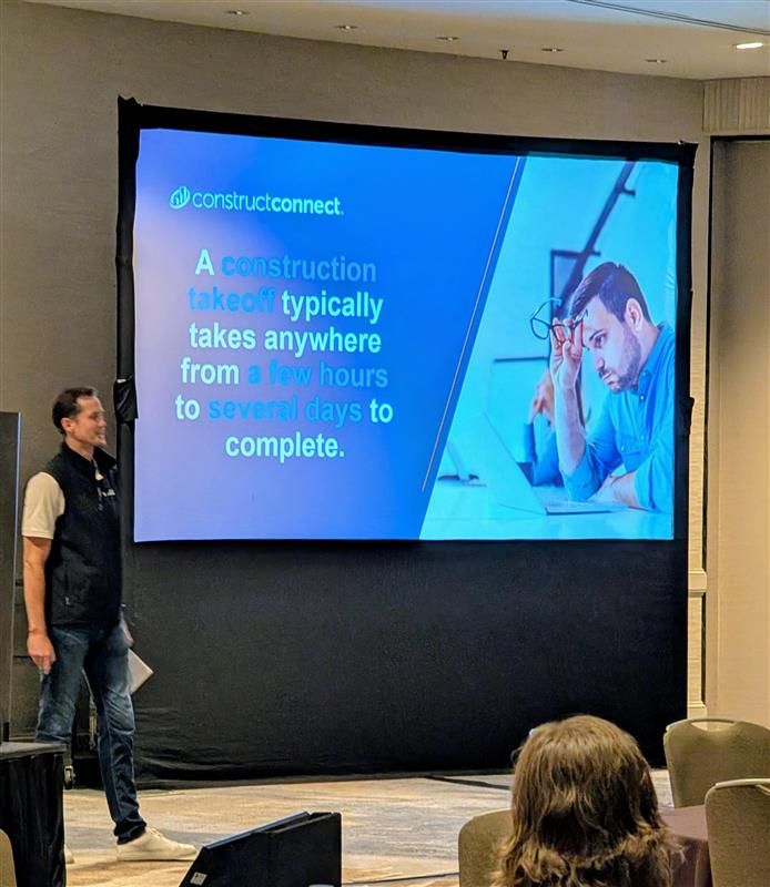 Theo Borsje gives a presentation in front of a large screen displaying the text, "A construction takeoff typically takes anywhere from a few hours to several days to complete," along with an image of a man working on a laptop, appearing thoughtful. The setting is a conference room.