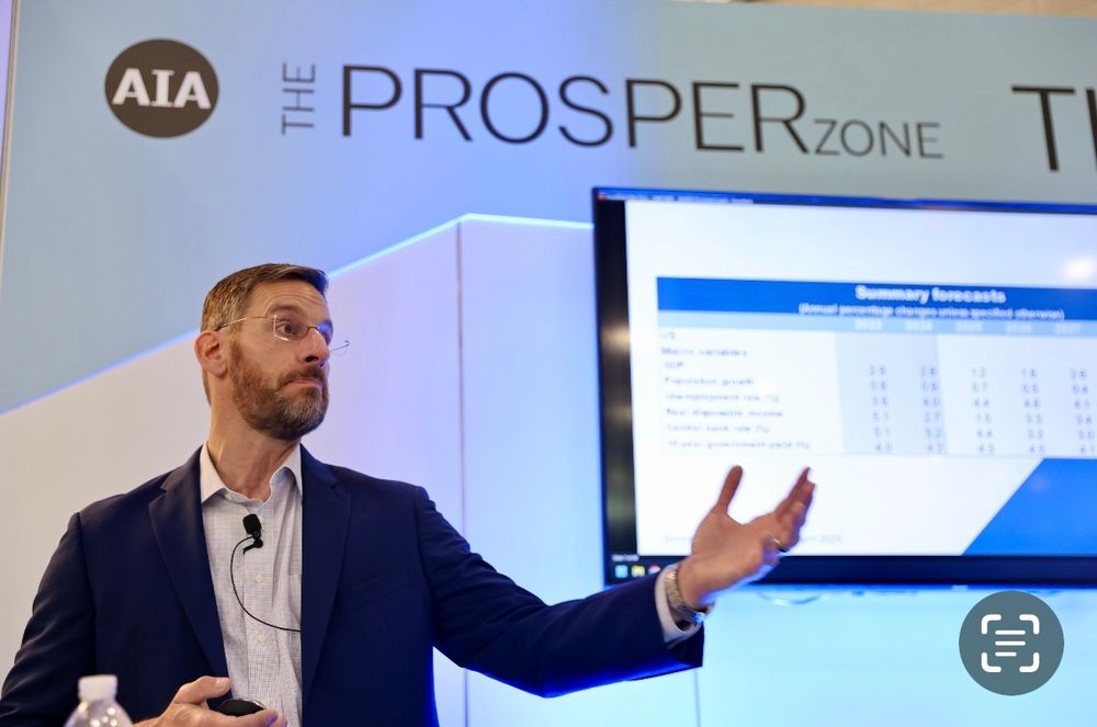 Man with glasses and a beard presents in front of a screen displaying a "Summary forecasts" table. The background shows a sign for "THE PROSPER Zone" and "AIA." He gestures with one hand, wearing a suit and a microphone. A water bottle is visible in the foreground.