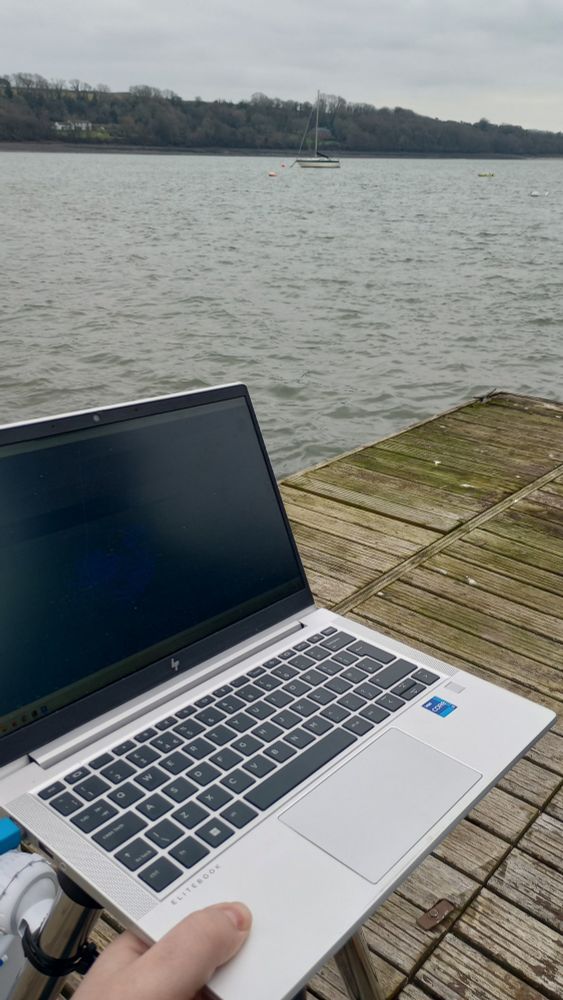 One laptop balanced on top of a tripod, with the river and moored boats in the background. The green/yellow boat from the previous image is just about visible in the background.