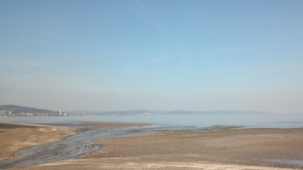 A view across Swansea bay. A stream runs across the sand from the bottom left of the image towards the sea. Swansea is on the left, with Baglan, Aberafan and Port Talbot and the hills behind them
