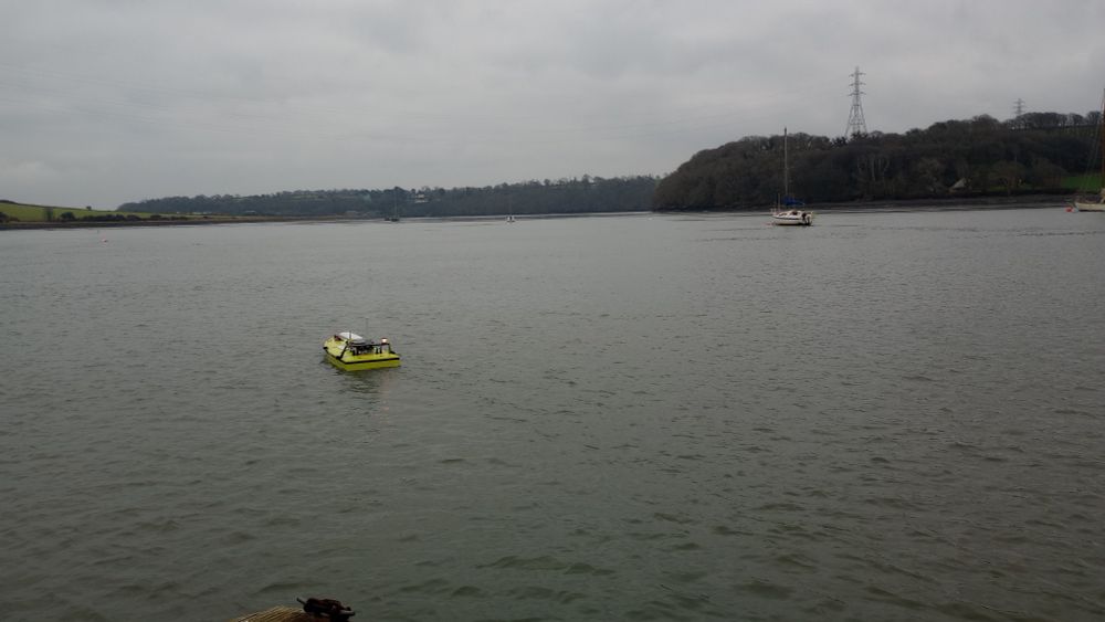 A small green/yellow boat with a white light on the rear and several antennae on top, floating one fairly calm looking river.