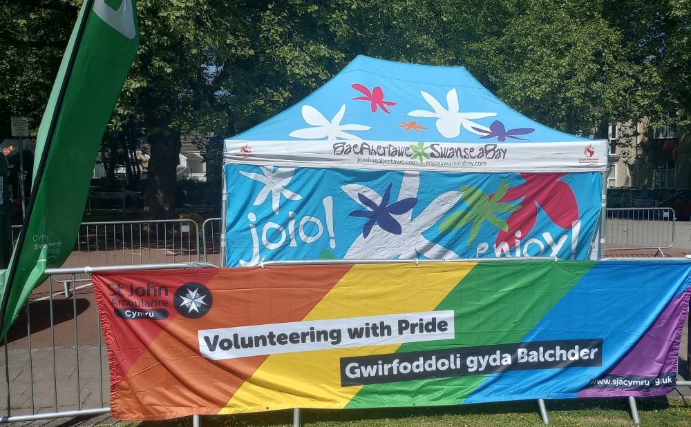 A blue tent with a St John Ambulance Cymru "Volunteering with Pride / Gwirfoddoli gyda Balchder" banner in front.