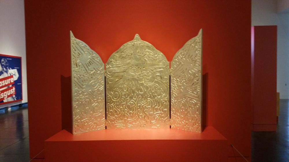 A full image of Altar Piece by Keith Haring, a golden triptych on a red display 