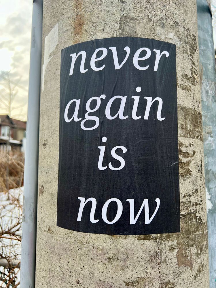 A concrete hydro pole with a black paper pasted to it that says ‘never again is now’ in white 