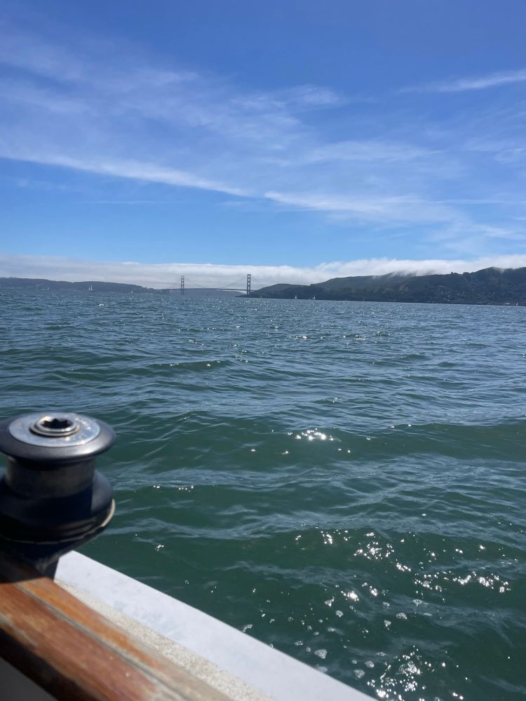 A view of the Golden Gate Bridge in the distance, over the rail of a small wooden sailboat. 
