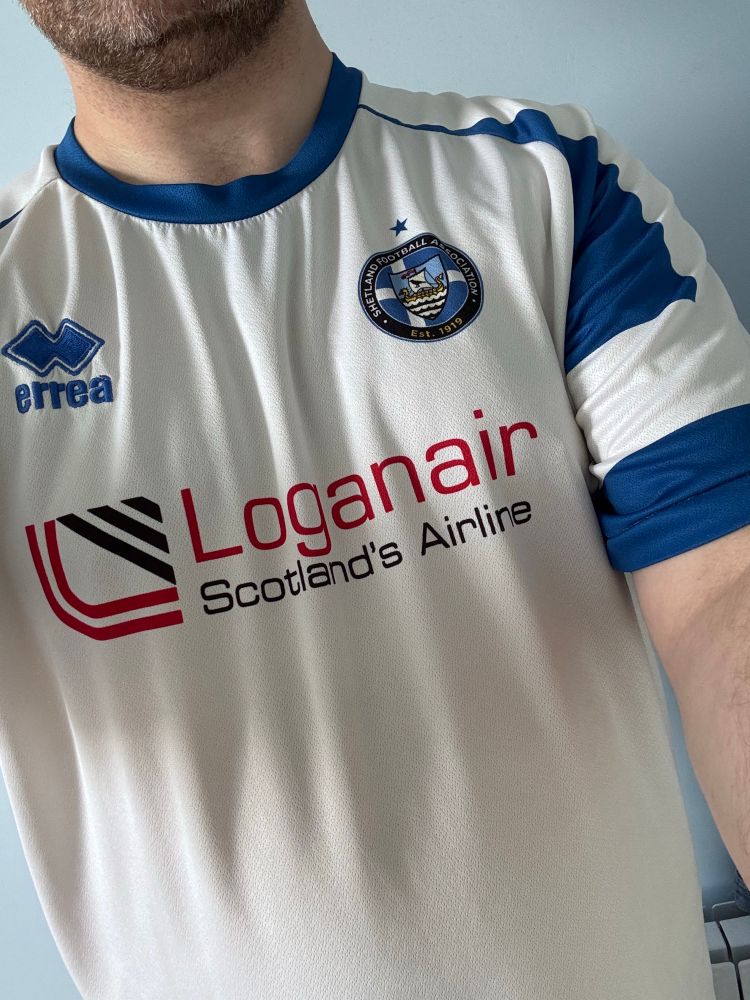 Torso of a man wearing a white Shetland Football Association top with blue and white sleeves in the pattern of the Shetland flag, kit made by Errea and sponsored by Loganair Scotland’s Airline.