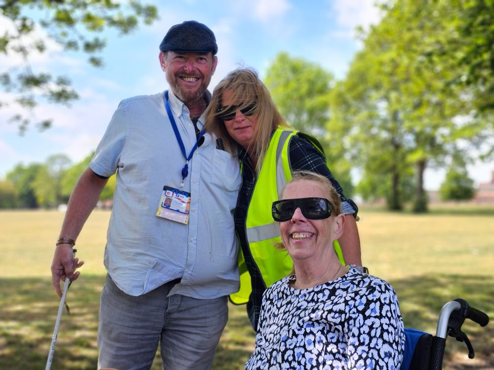 A group of three people huddled together for a photograph in a park. The man on the right is wearing a blue shirt, the woman in the middle is wearing a high vis vest, and the lady on the right of the image is sat in a wheelchair wearing dark sunglasses.