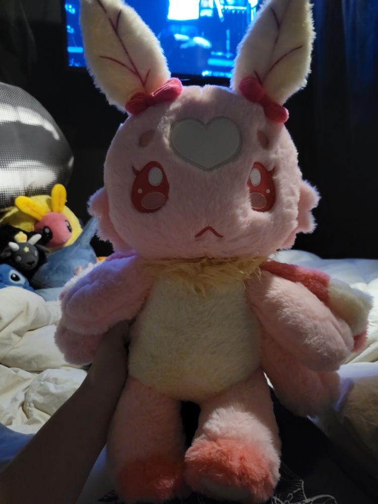 The world's cutest moth plushie. It's pink with a white belly and antenna. There are white hearts on the bottom feet and forehead and a small yellow ruff around the neck. In the background can be seen other plushies (mostly squishables) and a pillow of a mouth agape with vampire fangs that I got from Numbskullz at TSC - the shop collective. 