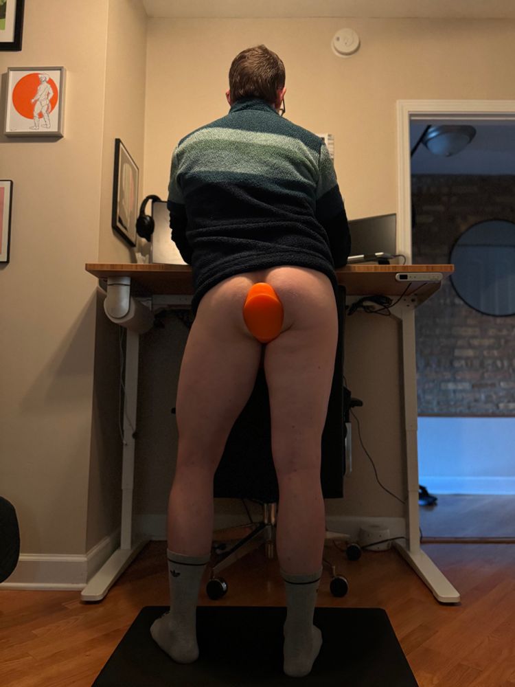 I’m standing at my desk while working from home pantless in a blue and green pullover and grey crew socks with a bright orange butt plug in my hole. 