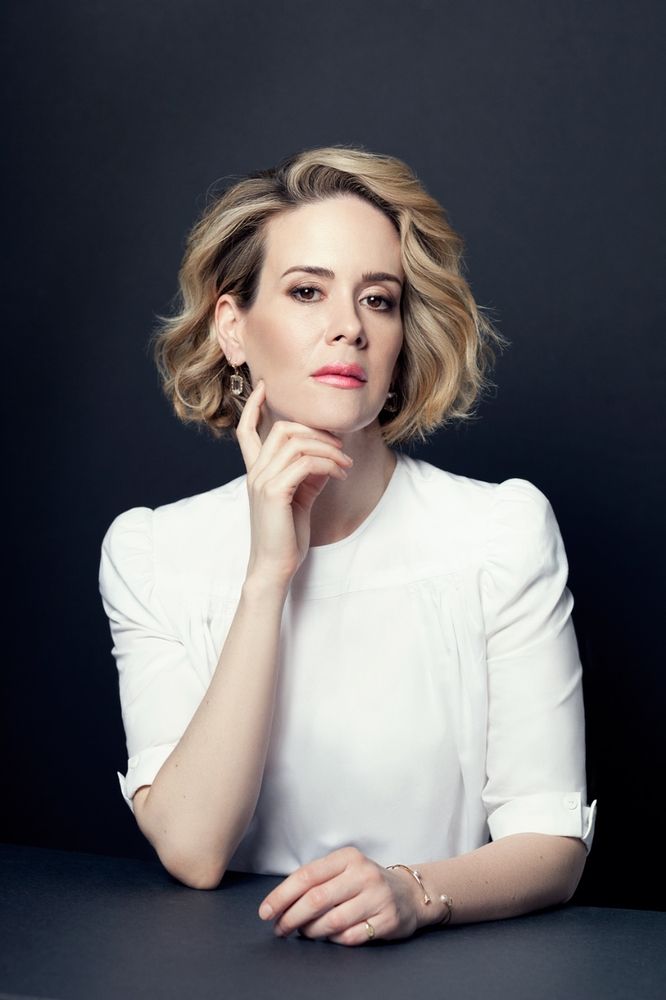 Image is a portrait of actress Sarah Paulson, who I find compelling as a performer, and who has a lisp.  

She wears a soft white blouse with cuffs the the sleeves, and shoulder length wavy blonde hair, posed against a black background, her left arm resting on a black table and the other hand at her chin. Her expression direct and thoughtful.