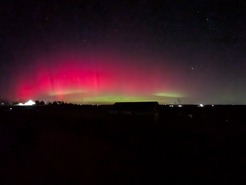 A zoomed-out view of the dark landscape. Green bands of aurora surrounded by a large red mass of aurora, with some stars also visible.