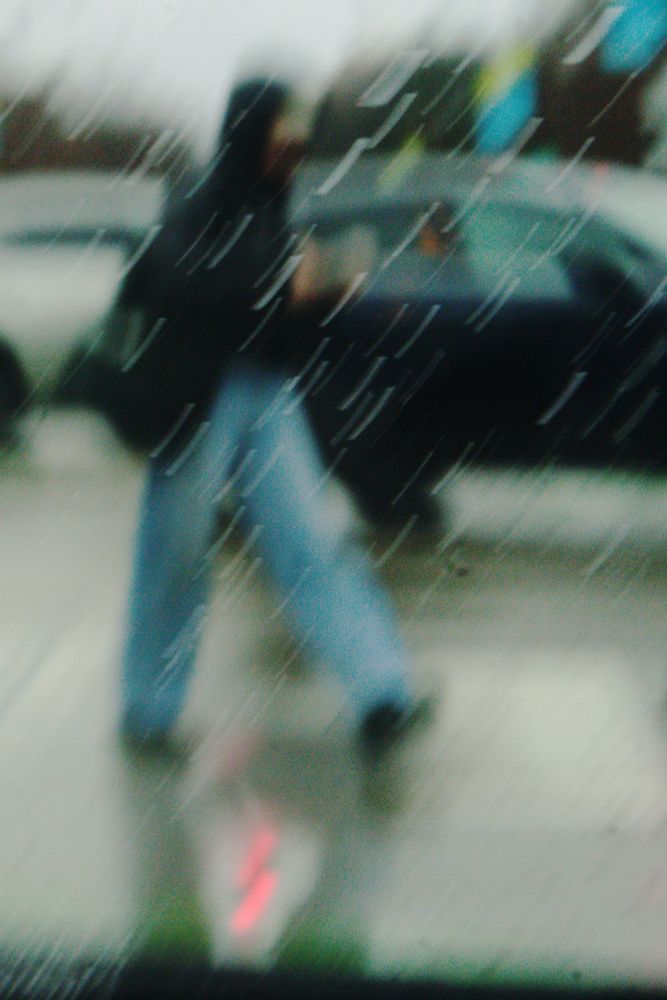 Photo of a pedestrian crossing a street in a snowstorm. She is blurry and out of focus, and the slow shutter speed makes the snow streak diagonally across the image like brush strokes, giving a dreamy, hazy quality to the image. Inexplicably, there is a neon reflection between her feet that looks like a red lightning bolt--presumably a reflection, but of something not in the rest of the frame.