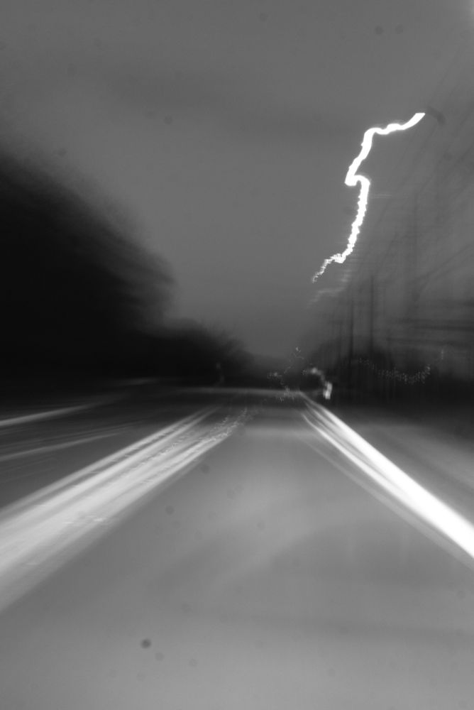 Black and white photo taken while driving down a two-lane highway. Looking straight down our lane toward the horizon. The camera is shaky, so all the lines are double or triple exposed with some blurriness. On the left are some silhouetted trees, in black. The pre-dawn sky is dark gray. On the right, we see some power lines in blurry outline. From the top right to the center of the image is an unexplained, lightning bolt-shaped weird white streak.