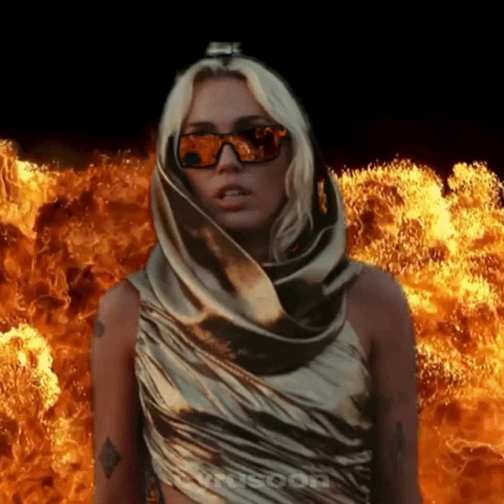 a woman wearing sunglasses and a scarf stands in front of flames