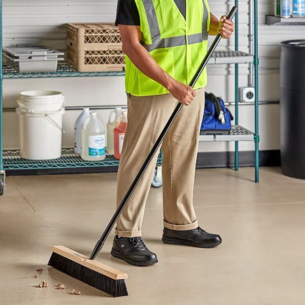 A shop or garage setting, with a person in a high visibility vest sweeping some debris with a good old fashioned push broom.