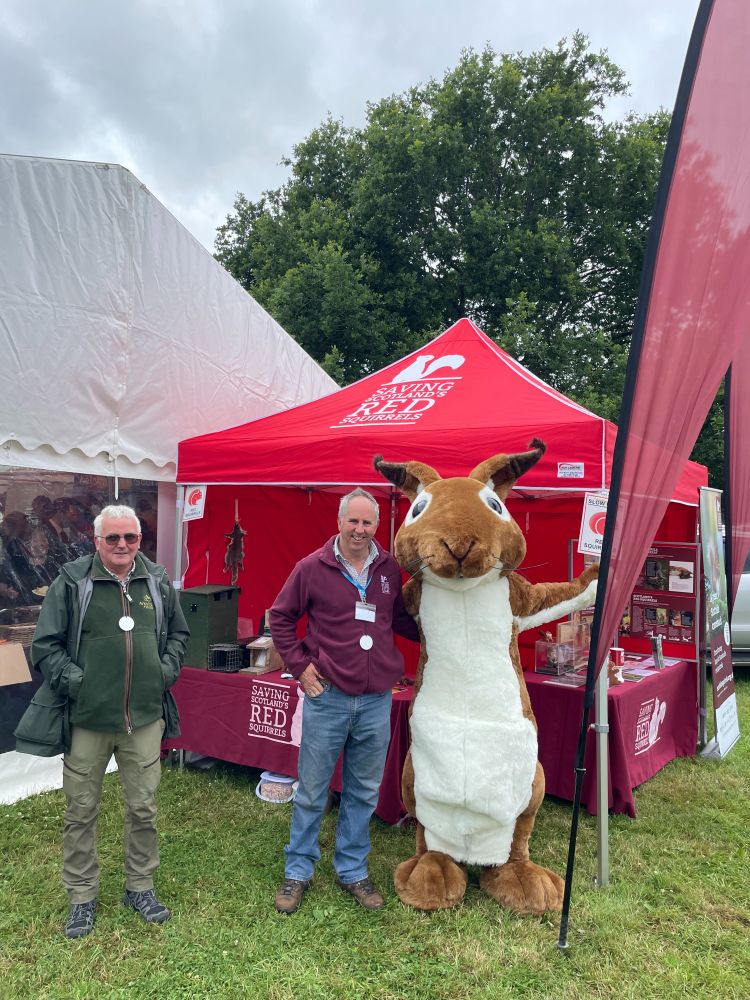 Members of the Saving Scotland’s Red Squirrels team are standing next to a table with branded materials, tablecloth and gazebo. They are wearing SSRS clothing and one person is wearing a giant red squirrel costume!
