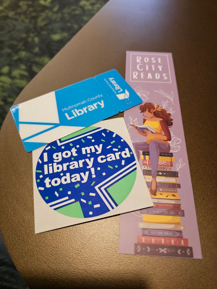 Blue and white plastic Multnomah County Library card. Blue, white, and green sticker that says "I got my library card today!" surrounded by confetti. Bookmark with "Rose City Reads" at the top and a drawing of a girl reading while sitting on top of a large stack of books.