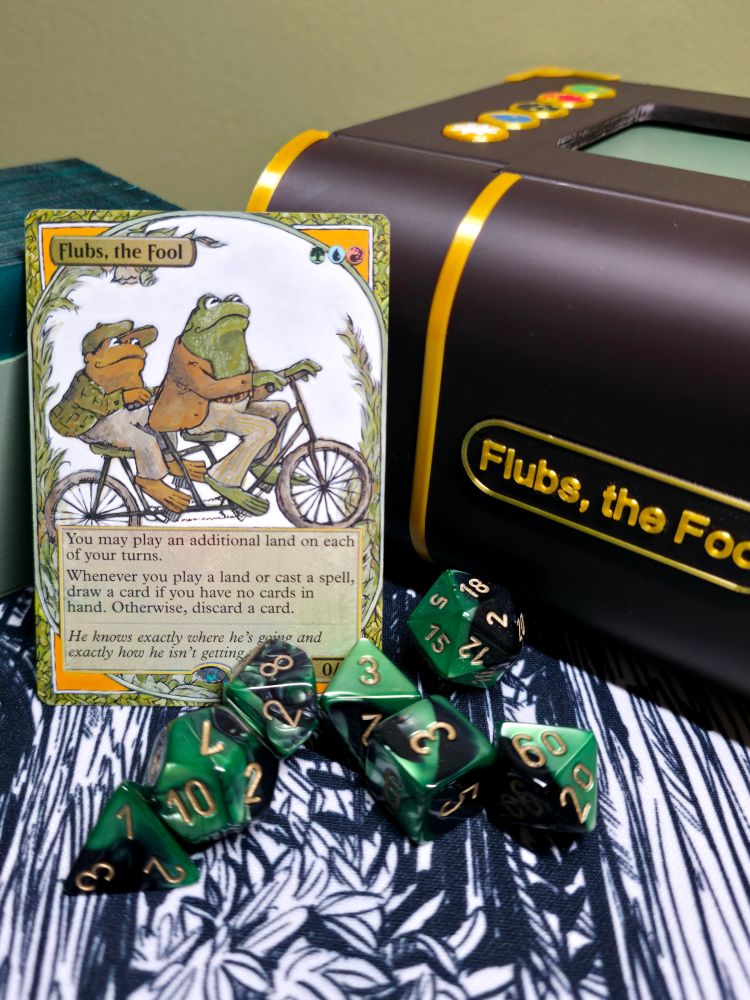 Magic the Gathering hand painted alteration by Baypaintstudios featuring Flubs the Fool as Frog and Toad. A Flubs the Fool deck box is in the background, with dice in the foreground. 
