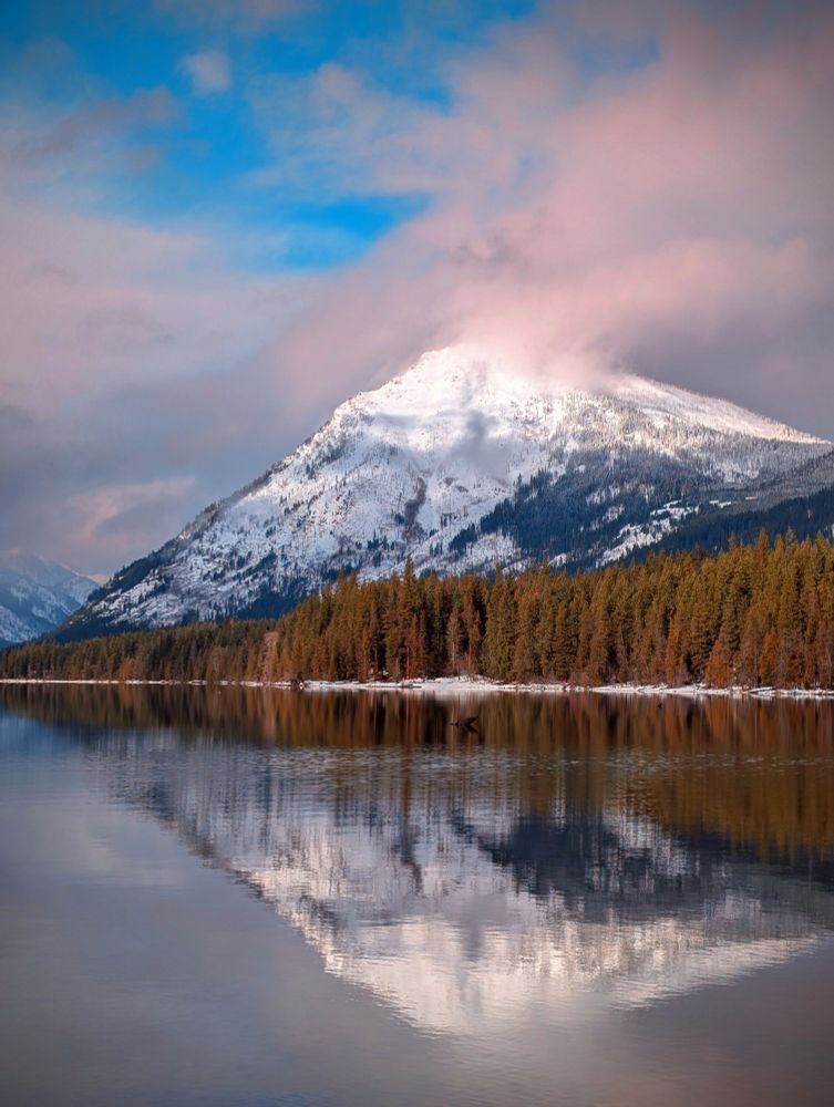 A photo across a lake of snow blowing off a mountain peak above a treeline in western Washington.