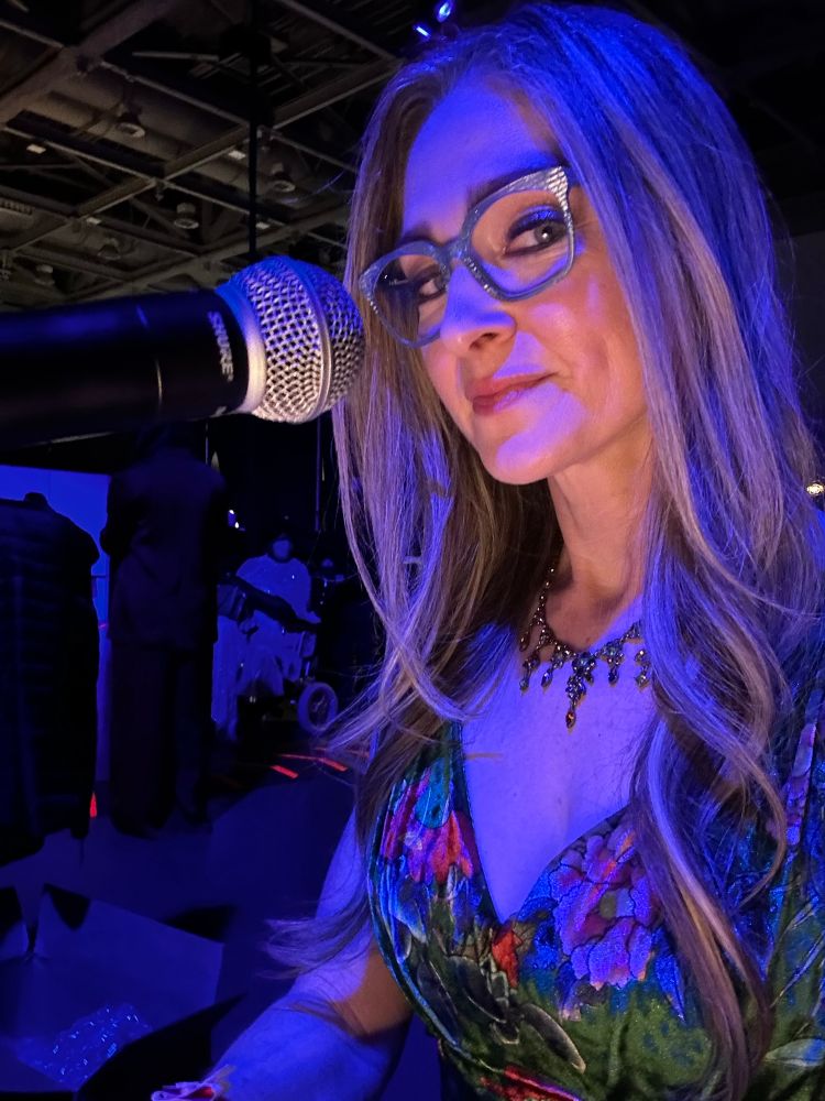 Julie is pictured sitting backstage at GDCA at a microphone wearing glasses and maybe a smirk. A friendly smirk. 