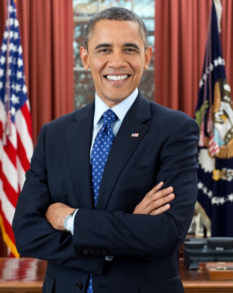 Presidential photo of Barack Obama in a dark suit, blue tie, smiling with arms crossed and an American flag in the background 