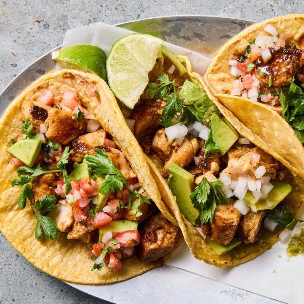 photo shows three soft chicken tacos on a plate and garnished with cilantro, avocado, and lime wedges 