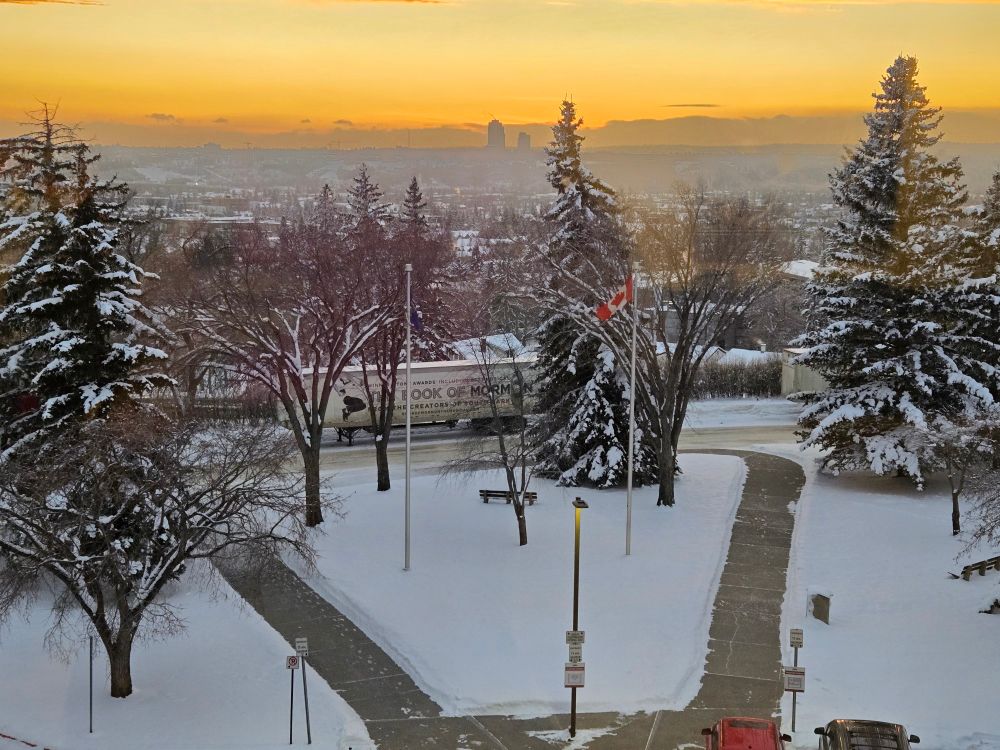 The Book of Morman musical truck at the end of a park Plaza with large snowy grass area, tall Spruce trees, raised Canada flag. In the background the city goes to fade with an orange winter sunset