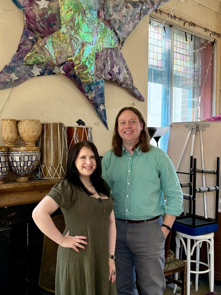A woman with long dark hair in an olive green dress and a man with a green checked shirt and grey chinos standing in an arts centre, surrounded by drums and various ephemera
