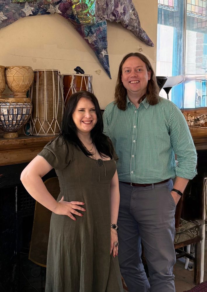 A woman with long dark hair in an olive dress with hand on her hip, and a man with long brown hair in a green shirt and grey chinos, with his hands in his pockets