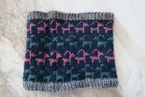 A navy cowl with grey rib and colourwork goats in pink and teal sits flat on a marble table.